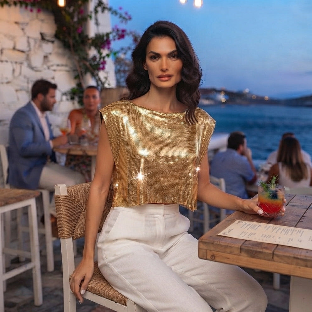 A woman seated at an outdoor seaside restaurant wears a shiny metallic gold sleeveless cropped top and white pants, holding a cocktail in a real environment with other diners and the ocean in the background.