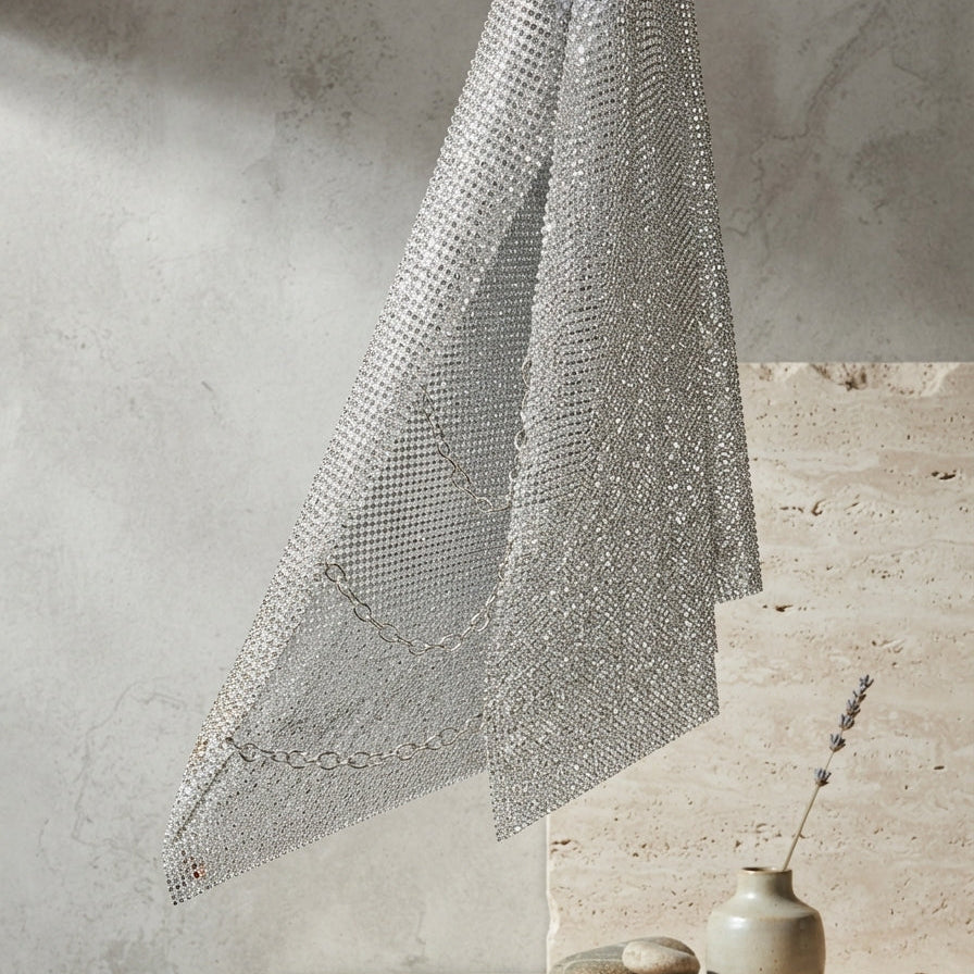 A silver metallic mesh fabric, likely a scarf or decorative textile, draped elegantly over a rustic stone shelf next to a small ceramic vase with dried lavender and smooth stones, set against a textured concrete wall.