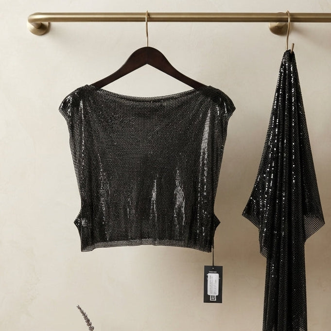 A black sequined cropped top and matching draped scarf hanging from a brass rod on a beige wall, with a small shelf and neutral decor below.