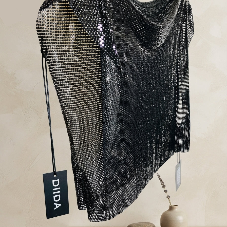 A black mesh chainmail-style top with fringe, held up by a hand against a beige wall. A black hangtag with the brand name 'DIIDA' is visible, and a rustic shelf with stones and a small vase sits below.